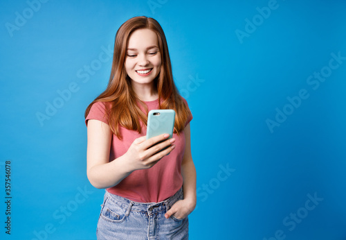 Young ginger girl in pink t-shirt and jeans posing isolated on blue background. Using mobile phone, typing sms message.
