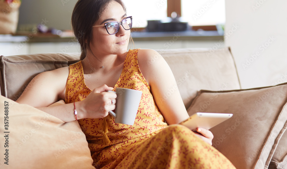 Young woman uses tablet while relaxing at home with coffee on the sofa couch - obrazy, fototapety, plakaty 