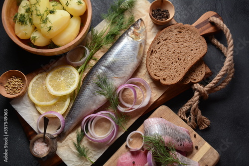 Herring fish on wooden board with pepper, herbs, red onion and lemon on black background. Top view with copy space with potatoes slices and rye bread