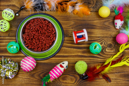 Set of toys for cat and bowl with dry pet food on wooden background. Top view. Pet care concept