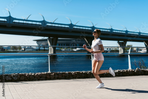Young fitness woman runner running in the morning on the promenade or embankment and doing sports exercises. Healthy lifestyle, street training, social distancing