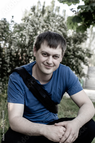 A man, 25, and 30, wearing a blue T-shirt with a backpack sits in the wild near trees.