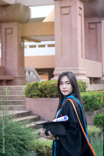 Portrait of a happy and excited young Asian female university graduate wears graduation gown and hat celebrates with a degree in the university campus on the commencement day. Education concept.