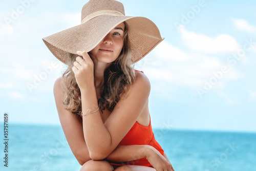 Beautiful young woman with long curly hair and a charming smile in a straw hat and a red bathing suit on the seashore, beach style and fashion, summer mood