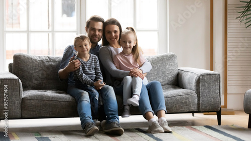 Family portrait cute adorable little kids sitting on mum and dad laps, looking at camera, smiling mother and father with preschool son and daughter posing for photo on cozy couch at home together