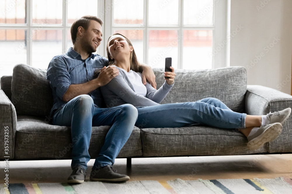 Happy young couple hugging, relaxing on couch, using smartphone, having fun with mobile device apps, smiling woman and man cuddling, enjoying leisure time, lazy weekend in living room at home Happy young couple hugging, relaxing on couch, using smartphone, having fun with mobile device apps, smiling woman and man cuddling, enjoying leisure time, lazy weekend in living room at home