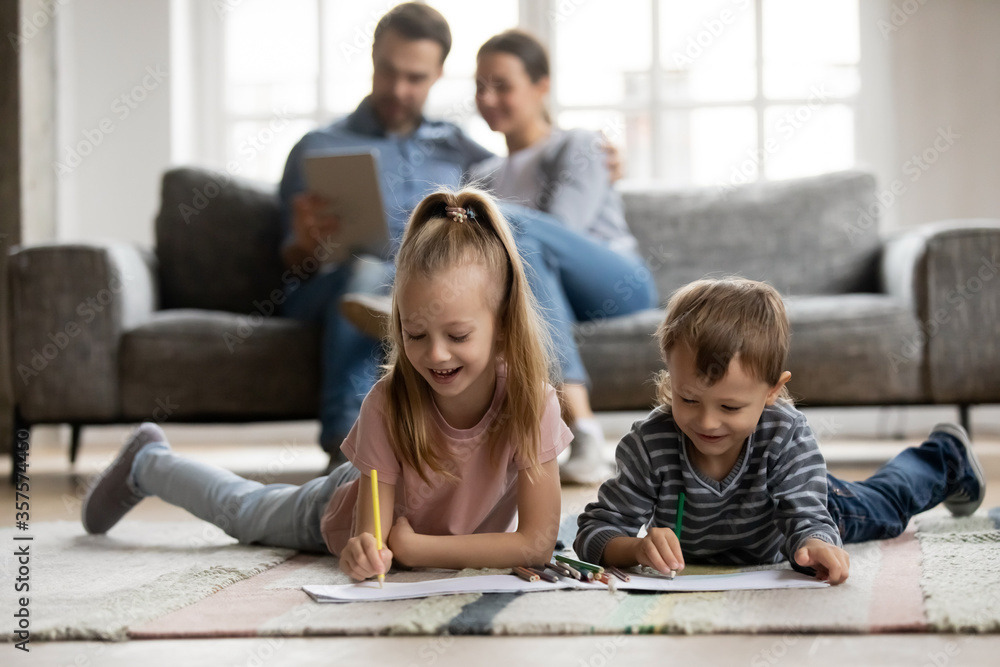 Little sister and brother playing on warm floor with underfloor heating while parents relaxing on couch in living room, cute girl and boy drawing with colorful pencils, enjoying leisure time - obrazy, fototapety, plakaty 