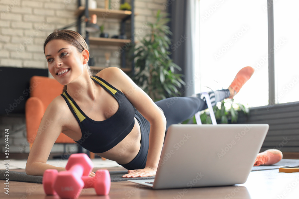 Fitness beautiful slim woman doing side plank with an elastic band and watching online tutorials on laptop, training in living room. Stay at home activities. Fitness beautiful slim woman doing side plank with an elastic band and watching online tutorials on laptop, training in living room. Stay at home activities.