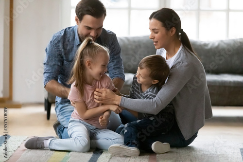 Happy young family playing funny game, sitting on warm floor with underfloor heating in living, laughing mother and father tickling little daughter and son, enjoying leisure time at home together