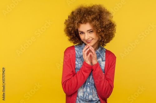 I have tricky plan! Portrait of devious cunning woman with fluffy curly hair clasping hands and smirking mysteriously, scheming cheats, evil prank. indoor studio shot isolated on yellow background