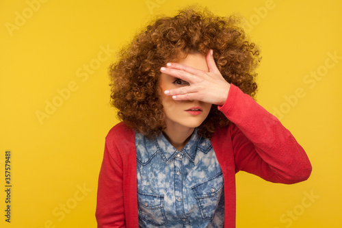 Portrait of curious nosy woman with curly hair spying rumors, looking through fingers with inquisitive expression, shy and scared to watch secret. indoor studio shot isolated on yellow background photo