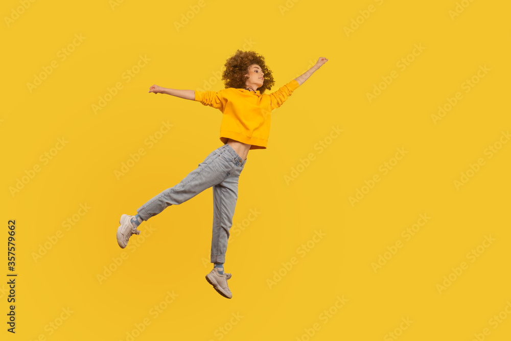 Portrait of superhero curly-haired girl in urban style outfit flying with raised hand high in air, feeling superpower and inspiration, achieving goal. indoor studio shot isolated on yellow background