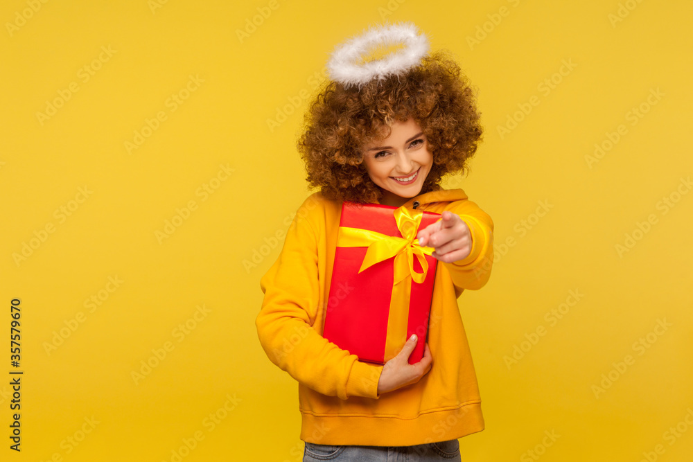Hey you, happy holidays! Portrait of cheerful curly-haired angelic woman with saint nimbus pointing to camera and holding wrapped christmas gift box. indoor studio shot isolated on yellow background Hey you, happy holidays! Portrait of cheerful curly-haired angelic woman with saint nimbus pointing to camera and holding wrapped christmas gift box. indoor studio shot isolated on yellow background