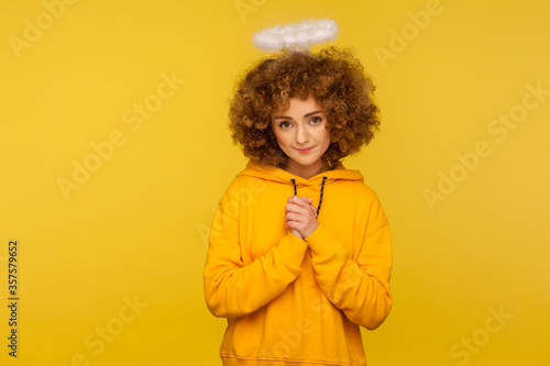 Portrait of kind lovely curly-haired hipster woman with saint nimbus holding hands in prayer and looking at camera with obedient modest expression. indoor studio shot isolated on yellow background