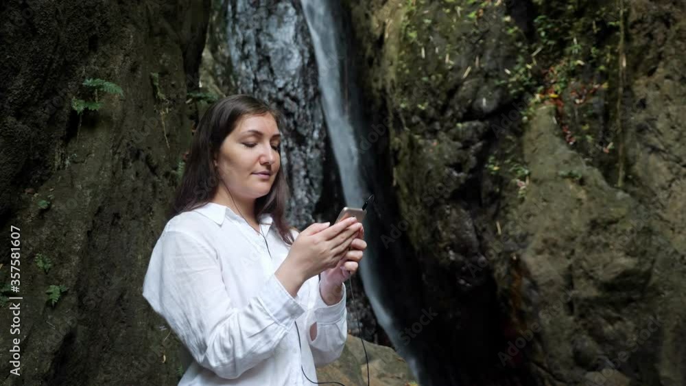 concentrated dark haired lady in headphones holds golden smartphone scrolls and reads information against tropical waterfall in reserve park close view concentrated dark haired lady in headphones holds golden smartphone scrolls and reads information against tropical waterfall in reserve park close view