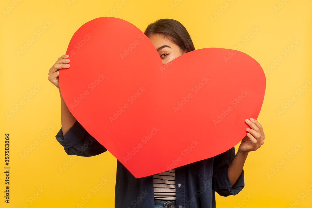 Portrait of happy playful girl peeking out of big read heart with alluring seductive look, flirting and expressing love, greeting on Valentine's day. indoor studio shot isolated on yellow background Portrait of happy playful girl peeking out of big read heart with alluring seductive look, flirting and expressing love, greeting on Valentine's day. indoor studio shot isolated on yellow background