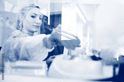 A young female scientist works in a sterile genetic or bacteriological laboratory with a dispenser and reagents in a medical gown, gloves and a doctor's cap.