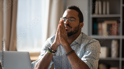 African guy sit at desk with clasped palms closed eyes thinking search problem solution, collect thoughts together, organize his emotional state before important meeting. Prayer hands sign of ask help photo