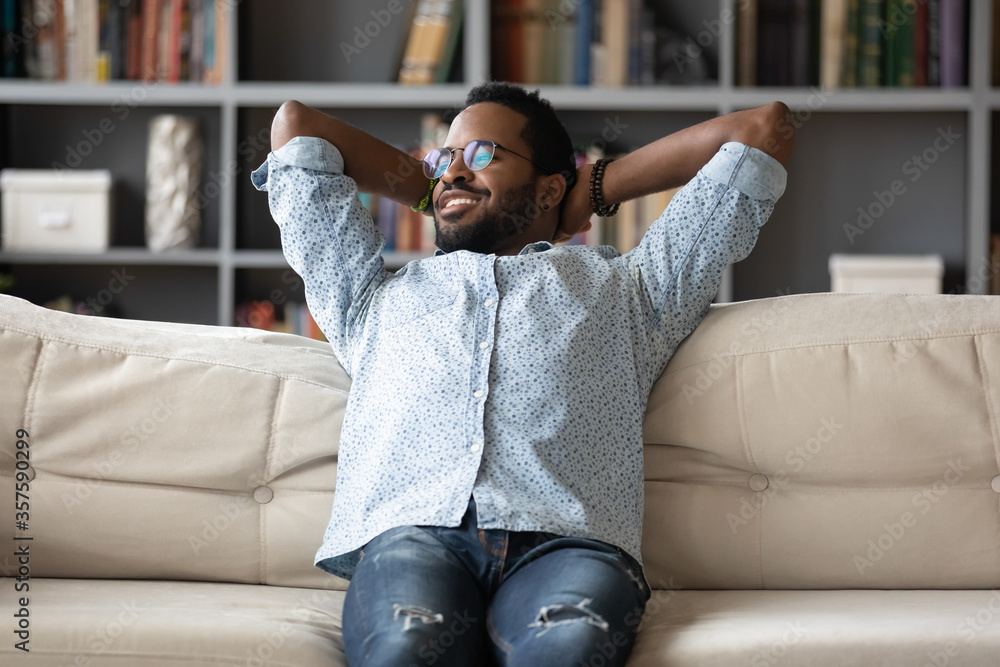 Obraz premium Peaceful African young man leaned on comfortable couch putting hands behind head relaxing looking out the window enjoy lazy weekend, has positive thoughts fresh conditioned air at modern home concept