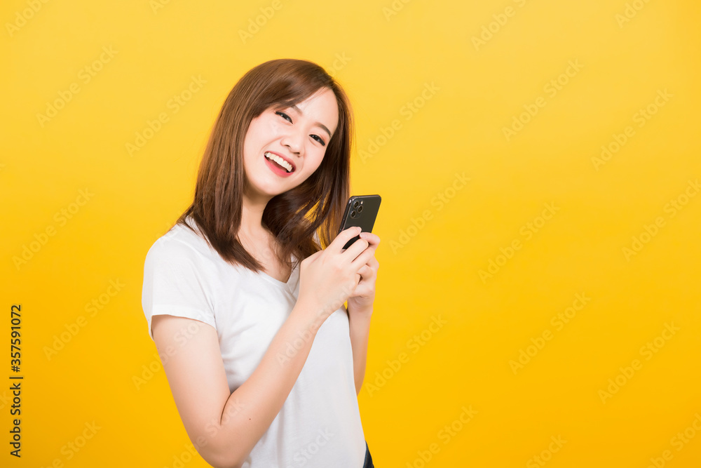 Asian happy portrait beautiful cute young woman teen smiling standing wear t-shirt using smart mobile phone looking to camera isolated, studio shot on yellow background with copy space Asian happy portrait beautiful cute young woman teen smiling standing wear t-shirt using smart mobile phone looking to camera isolated, studio shot on yellow background with copy space