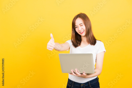 Asian happy portrait beautiful cute young woman teen smiling standing wear t-shirt using laptop computer and showing thumb up looking to camera isolated, studio shot yellow background with copy space