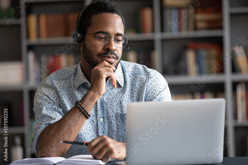 Focused African man wear headphones with microphone looking at laptop screen listens audio lesson learn foreign language with tutor makes video call. Student watching webinar, e study on-line concept