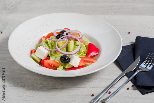 White plate with vegetable salad "Greek" with feta cheese (Ingredients: tomatoes, fresh cucumbers, red onions, bell peppers, olives, feta cheese and Greek dressing) on a light gray background with dar