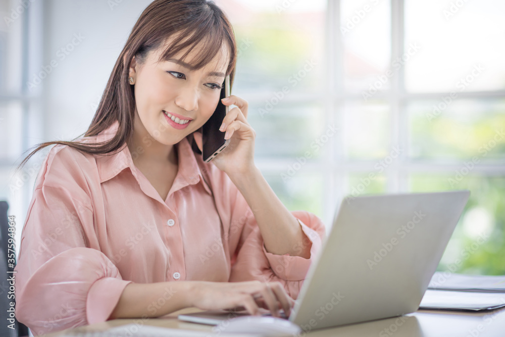 Beautiful Asian woman is talking on the phone in the office. With laptop placed on the table and typing on keyboard. Asian business people, employee, and office staff. Concept Beautiful Asian woman is talking on the phone in the office. With laptop placed on the table and typing on keyboard. Asian business people, employee, and office staff. Concept
