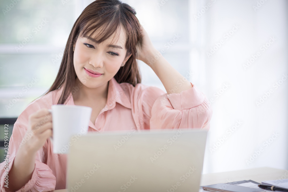 Beautiful Asian woman with long hair Wearing a pink shirt holding a white coffee cup and looking at the laptop. Relax and drinking coffee in office, coffee shop, coffee break concept. Beautiful Asian woman with long hair Wearing a pink shirt holding a white coffee cup and looking at the laptop. Relax and drinking coffee in office, coffee shop, coffee break concept.