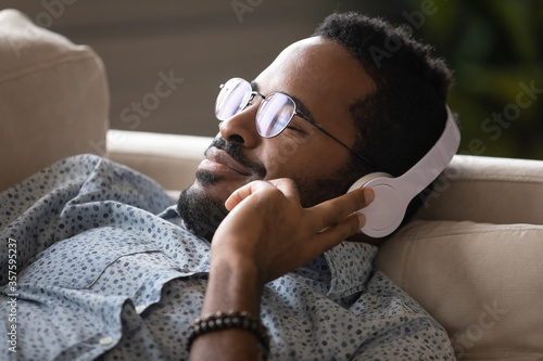 Close up african calm guy in headphones relaxing lying on couch in living room at home enjoy pause carefree time and inner balance, no stress, listening favorite music, e-book, pleasant sound concept