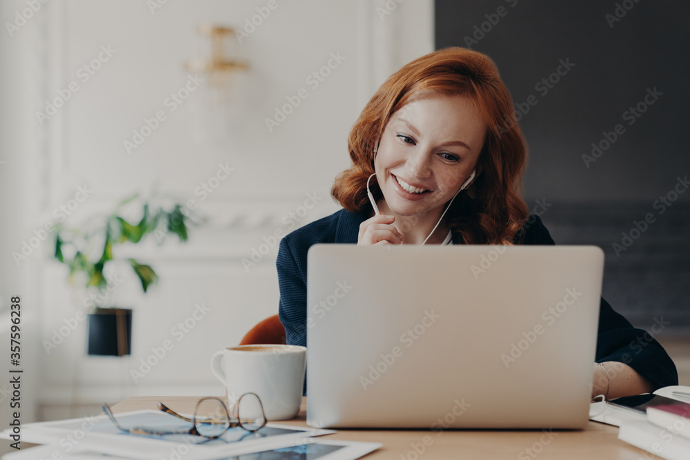 Positive ginger businesswoman works remotely from home during quarantine, has video conference with colleagues, makes online call, uses wireless internet and modern laptop computer with earphones Positive ginger businesswoman works remotely from home during quarantine, has video conference with colleagues, makes online call, uses wireless internet and modern laptop computer with earphones