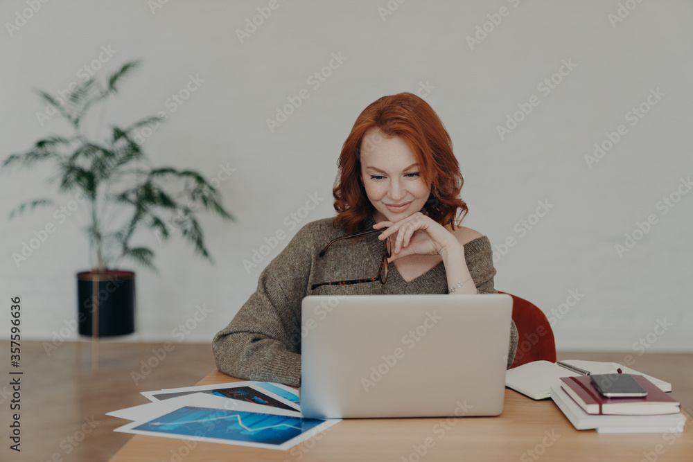 Busy woman freelancer holds spectacles for vision correction, poses at home desktop, has paperwork and works on laptop computer, concentrated at notebook screen. Technology and communication Busy woman freelancer holds spectacles for vision correction, poses at home desktop, has paperwork and works on laptop computer, concentrated at notebook screen. Technology and communication
