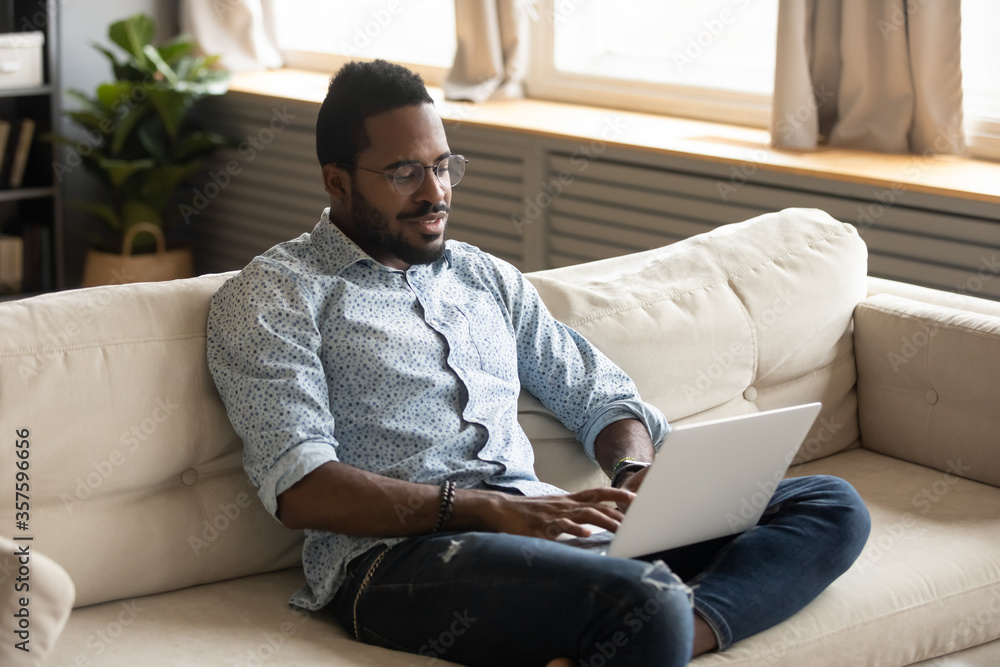 African guy in eyeglasses resting on comfy couch in cozy living room spend free time at home enjoy lazy weekend with pc have fun in internet, web surfing, search information, modern tech usage concept African guy in eyeglasses resting on comfy couch in cozy living room spend free time at home enjoy lazy weekend with pc have fun in internet, web surfing, search information, modern tech usage concept