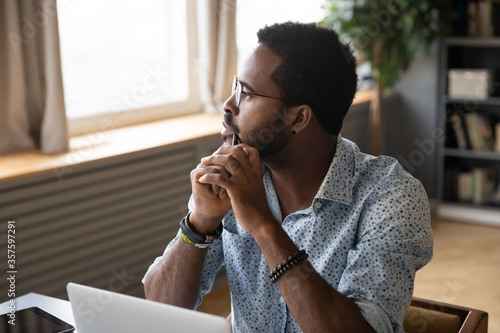 African guy sitting at table near laptop pondering over problem search answer looking at distance out the window. Student or office worker having doubts, feel uncertain thinking about solution concept