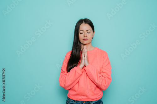 Peaceful young beautiful female dressed in casual, keeps hands in praying gesture, has calm expression, asking good luck, stands against bright turquoise background. Young brunette woman prays.