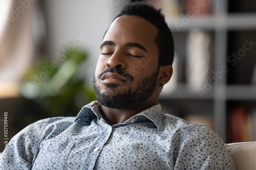 Close up African young man sit on couch closed eyes resting reduces fatigue, enjoy lazy day, breath fresh air at conditioned home, visualizing and meditating improve inner balance, no stress concept