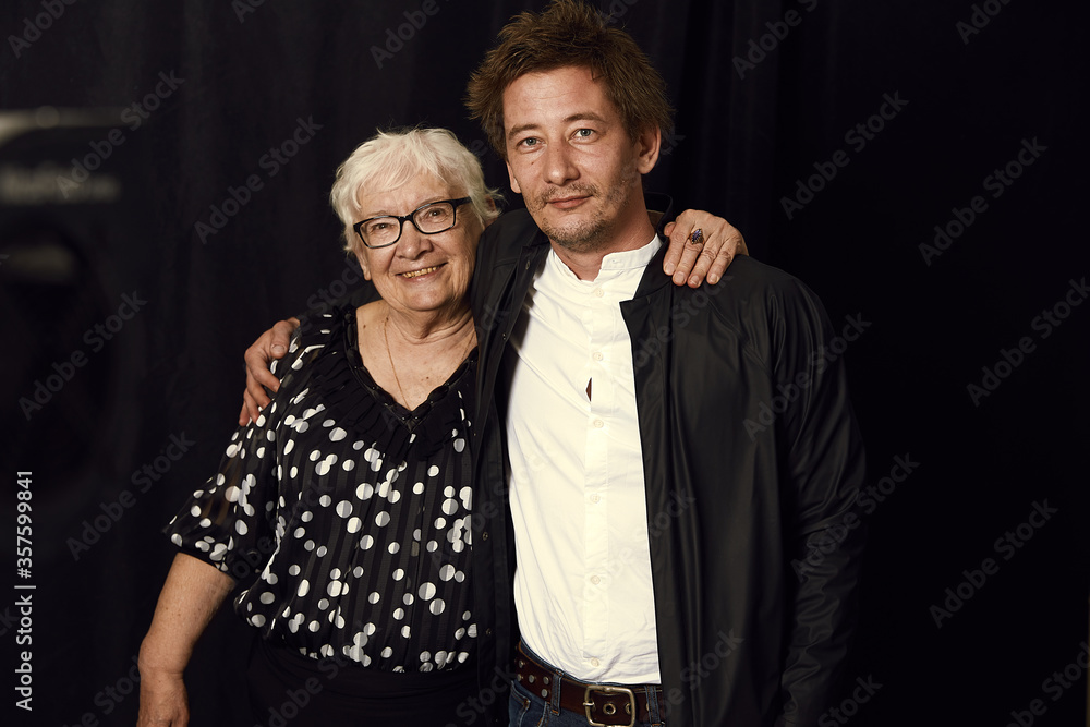 Two generations. Indoor family portrait of mature wrinkled gray haired woman dressed in stylish blouse, surrounded with love of son, enjoying time together, isolated on black studio wall. Film effect.