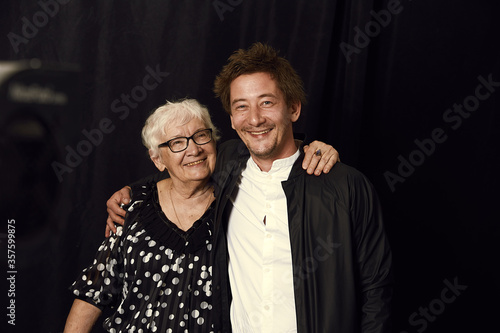 Two generations. Indoor family portrait of mature wrinkled gray haired woman dressed in stylish blouse, surrounded with love of son, enjoying time together, isolated on black studio wall. Film effect.