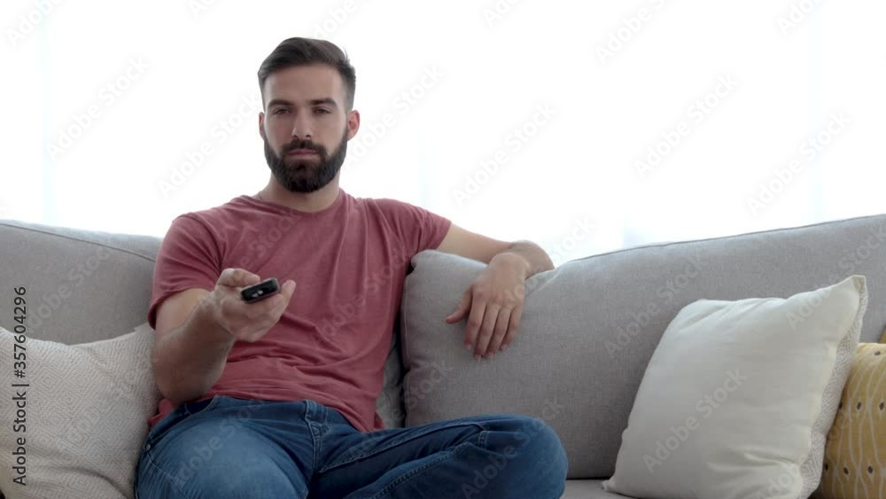 Young casual bearded man changing the channels on the tv or television remote control trying to find something interesting to watch on his day off or free day from his work Young casual bearded man changing the channels on the tv or television remote control trying to find something interesting to watch on his day off or free day from his work
