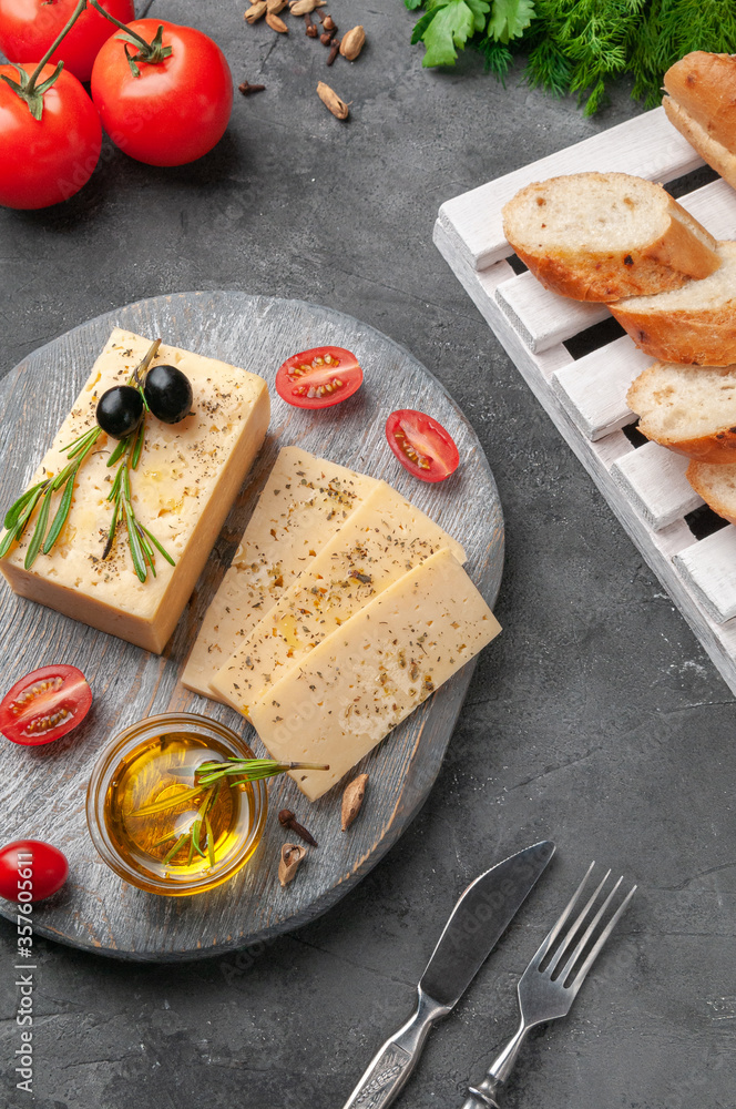 A piece of semi-hard cheese is sliced ​​into plastics. Lies on a round gray wooden board. Garnished with spices, rosemary and olives.  Gray concrete background. View from above.