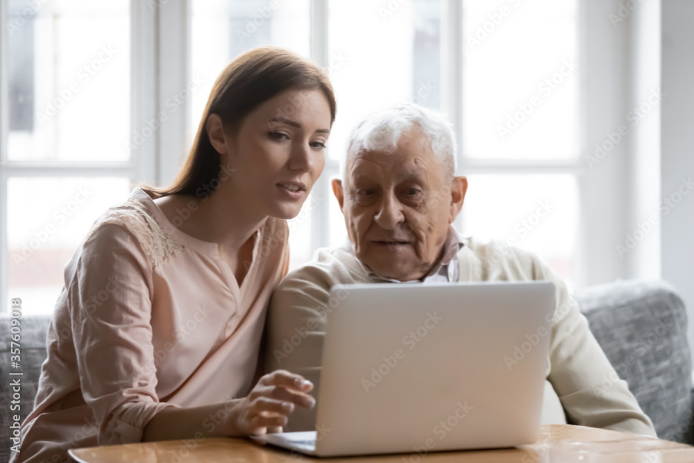 Caring grownup daughter teaching elderly father to use laptop, young woman and mature man looking at computer screen, sitting on couch at home, watching movie, making video call or shopping online - obrazy, fototapety, plakaty 
