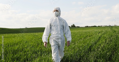 Caucasian male farmer ecologist in white protective costume and goggles walking in green field in summer. Man scientist and biologist strolling the margin with eco harvest.