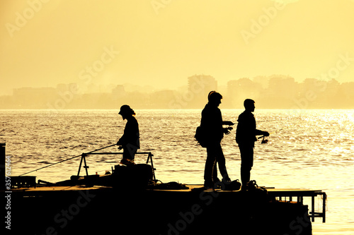 Silhouetted people relaxing and fishing on Antalya harbor over yellow colored dusk time in Antalya, Turkey