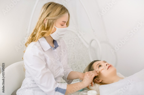 Beautiful young blond woman is getting face skin treatment. Female doctor cosmetologist is holding a bottle of cream and a spatula and applying cream on patient face