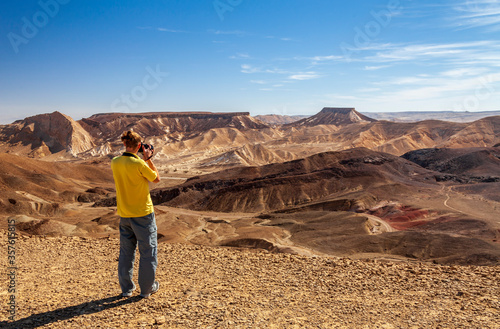 A young man on top of a mountain photographs a mountain landscape at the camera. Ramon Crater, Israel. Travel and photos concept.