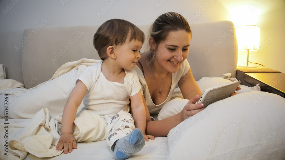 Happy smiling mother with toddler boy watching video on tablet at night