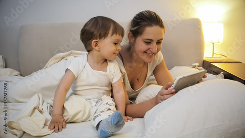 Happy smiling mother with toddler boy watching video on tablet at night