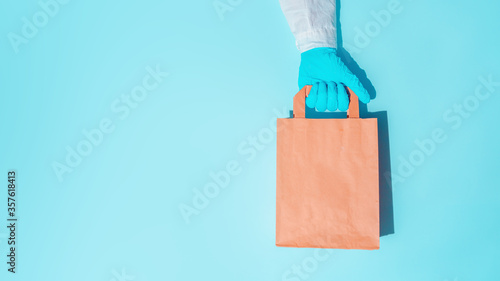 hand in gloves holding shopping paper bag. Concept of consumerism, shopping symbol during pandemic time. Copy space trend pastel color. Hard shadows, morning light.