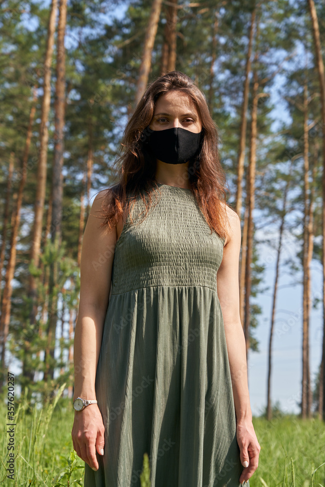 Young woman with a black mask walks outdoors in the forest at a sunny summer day.