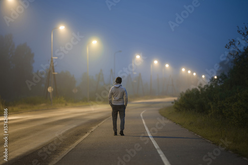 One young man slowly walking on long sidewalk under street lights in summer night. Spending time alone. Back view.
